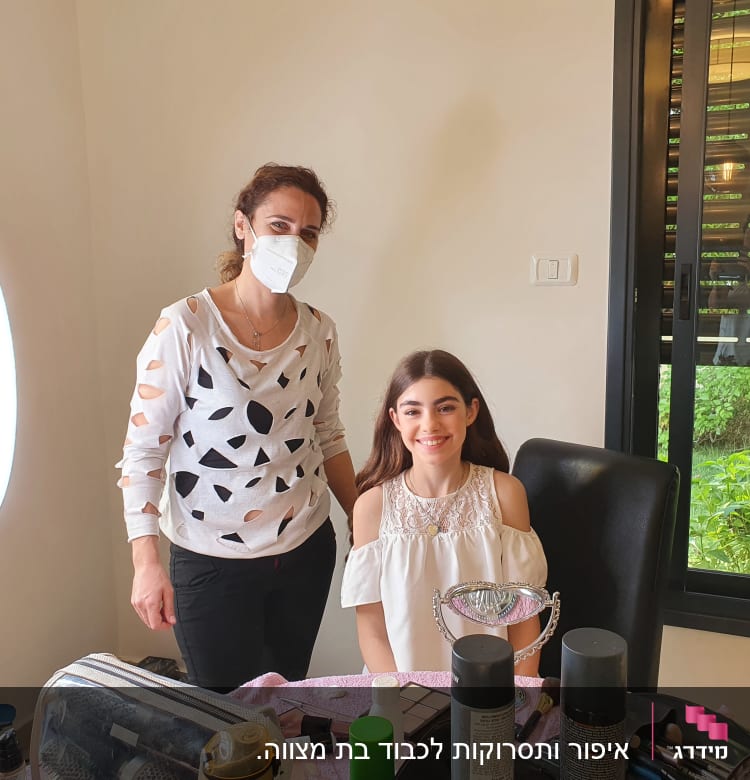 אישה עם מסכה ליד אישה צעירה עם איפור, מראה ומוצרי שיער על השולחן.